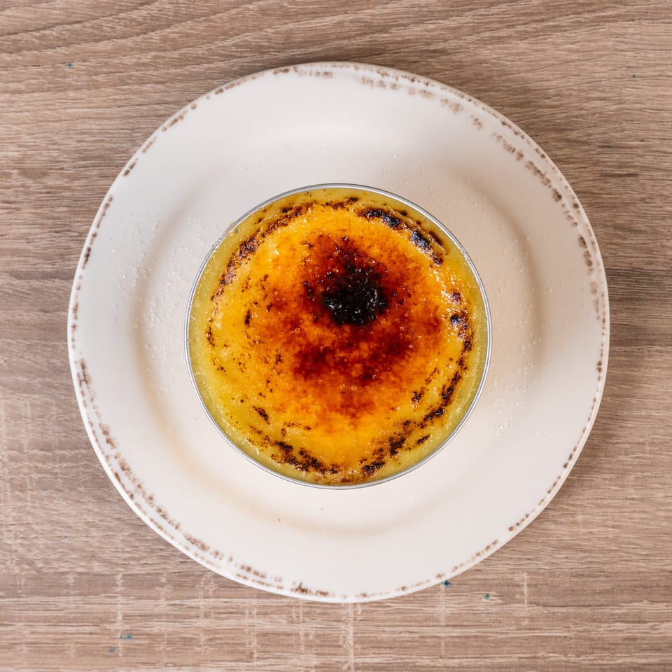 A crème brûlée with a caramelized sugar topping, served on a white plate on a wooden table.